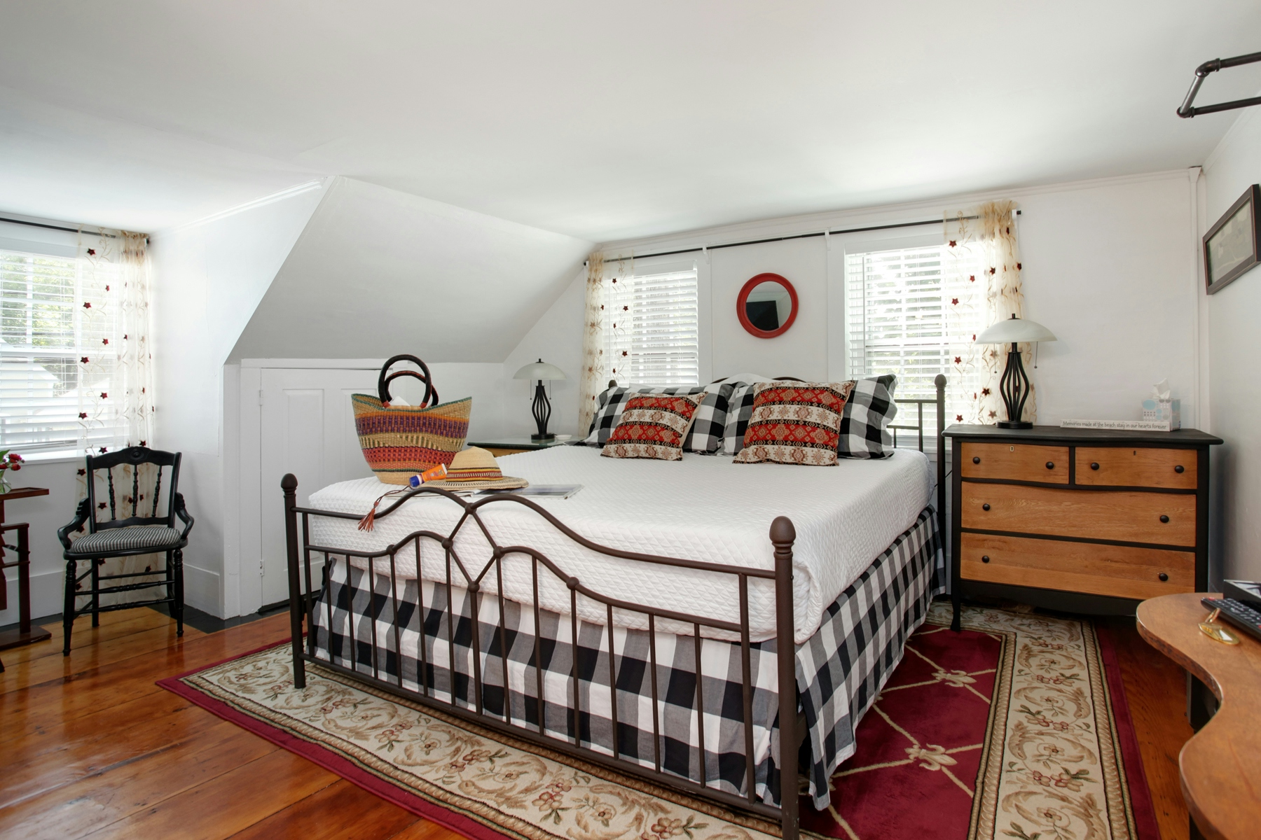 View of bed with black and white plaid bedding, natural wood and black painted bureau, and red and cream floral area rug