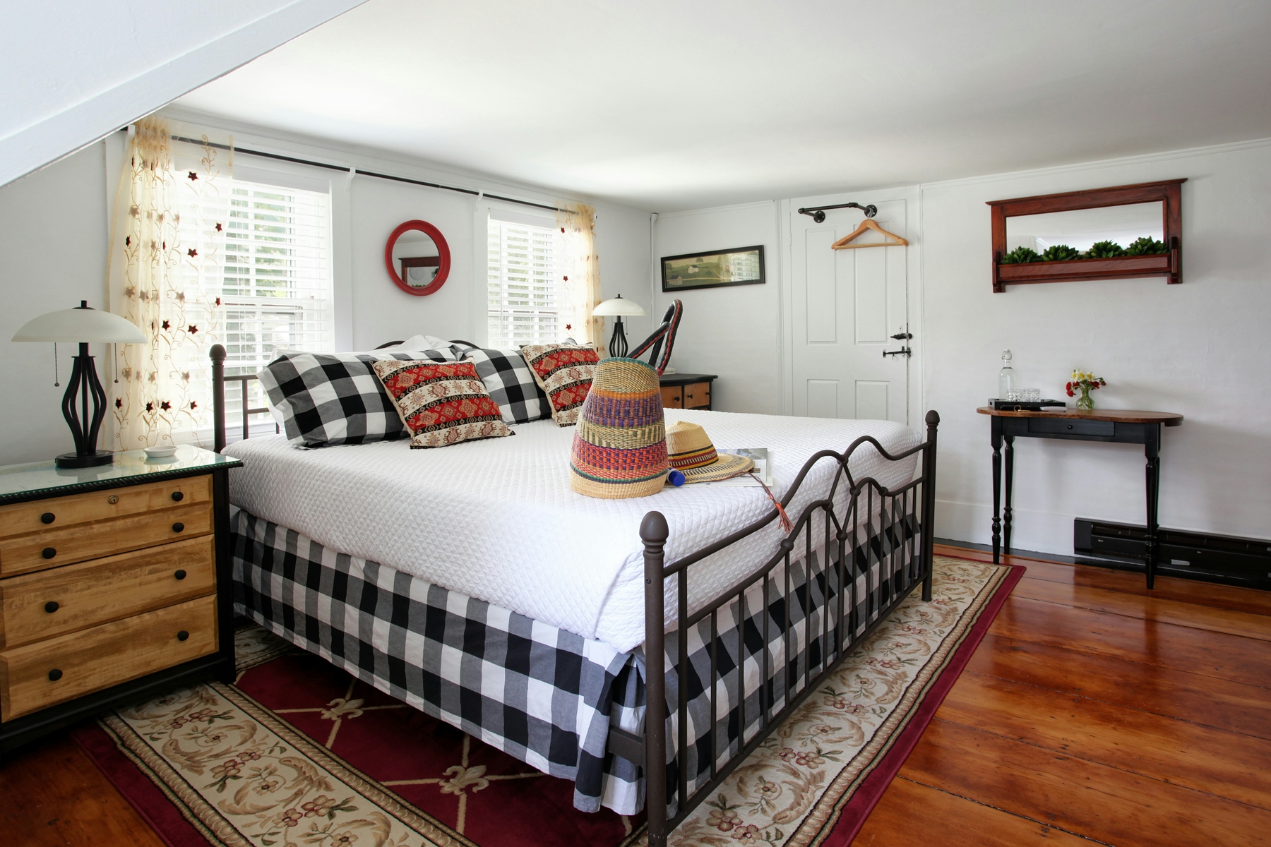 View of bed with black and white plaid bedding, natural wood and black painted bureau, finished wood floor with red area rug