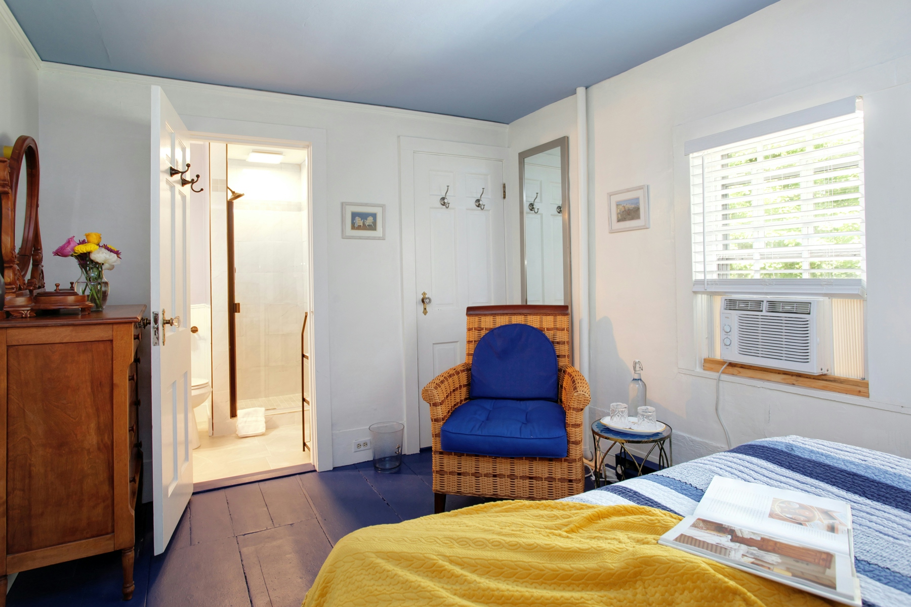 Area opposite from head of bed with cozy yellow and blue upholstered chair, window, bureau, and door leading into bathroom