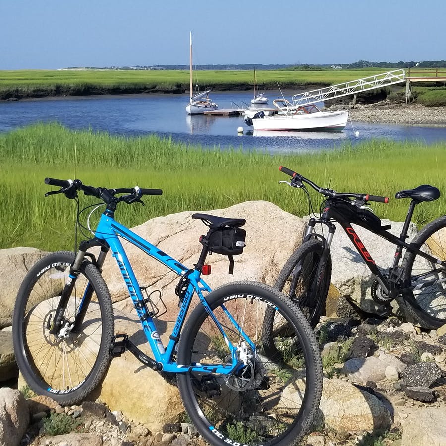 Two bikes, parked in gravel with big rocks separating the green marshy area to the blue water, where there is a dock and two small sail boats.