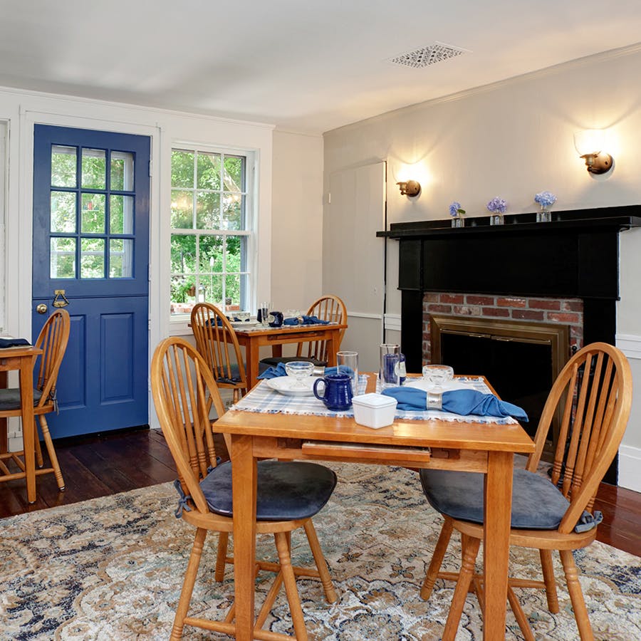Square Pine tables and chairs with blue linens and table settings, blue exterior door, and black fireplace with multi color rug. 