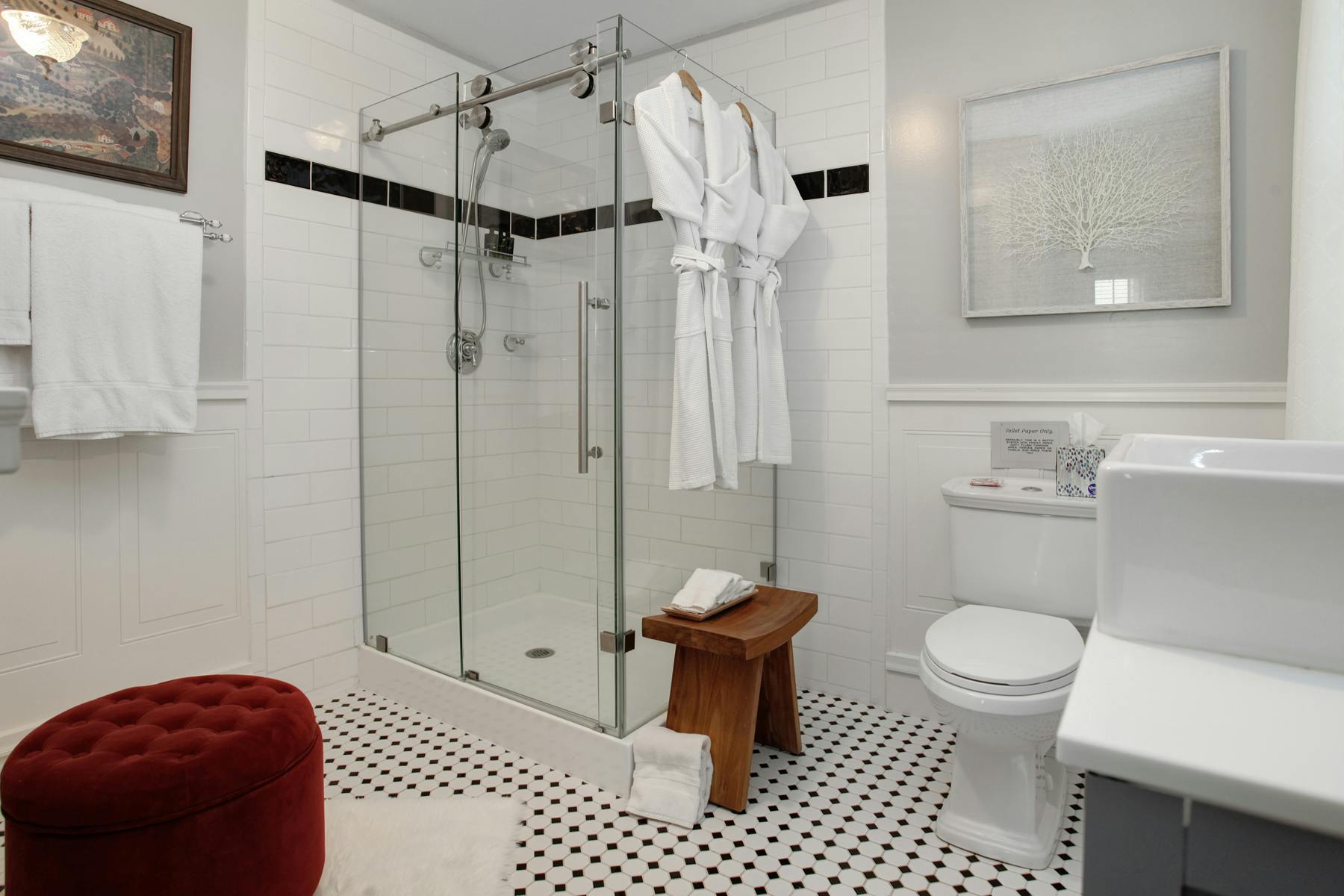 Bathroom with glass enclosed shower, toilet, and vanity, plush white towels and robes, black & white tiled floor and red upholstered ottoman