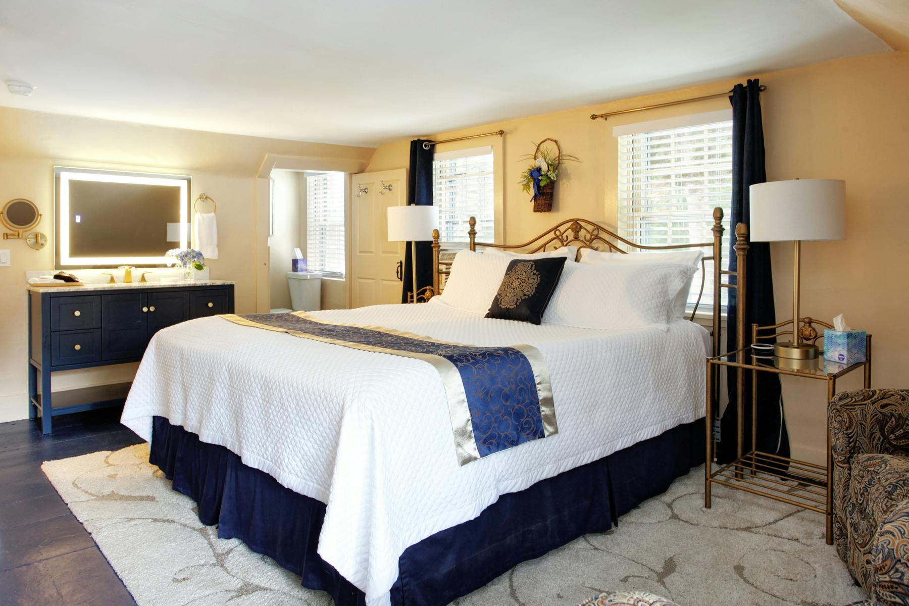 View of bed with white and navy bedding, end tables with lamps, wooden navy bureau with mirror, and neutral plush area rug