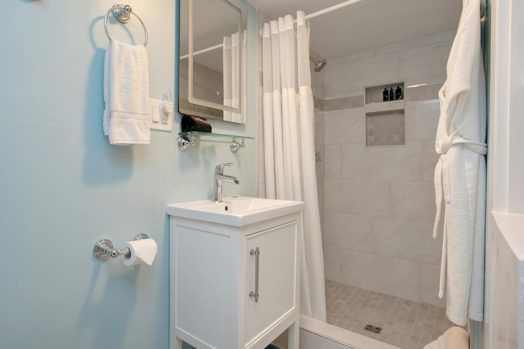 View of bathroom with white vanity and sink, shower, and white plush towels and robe hanging up