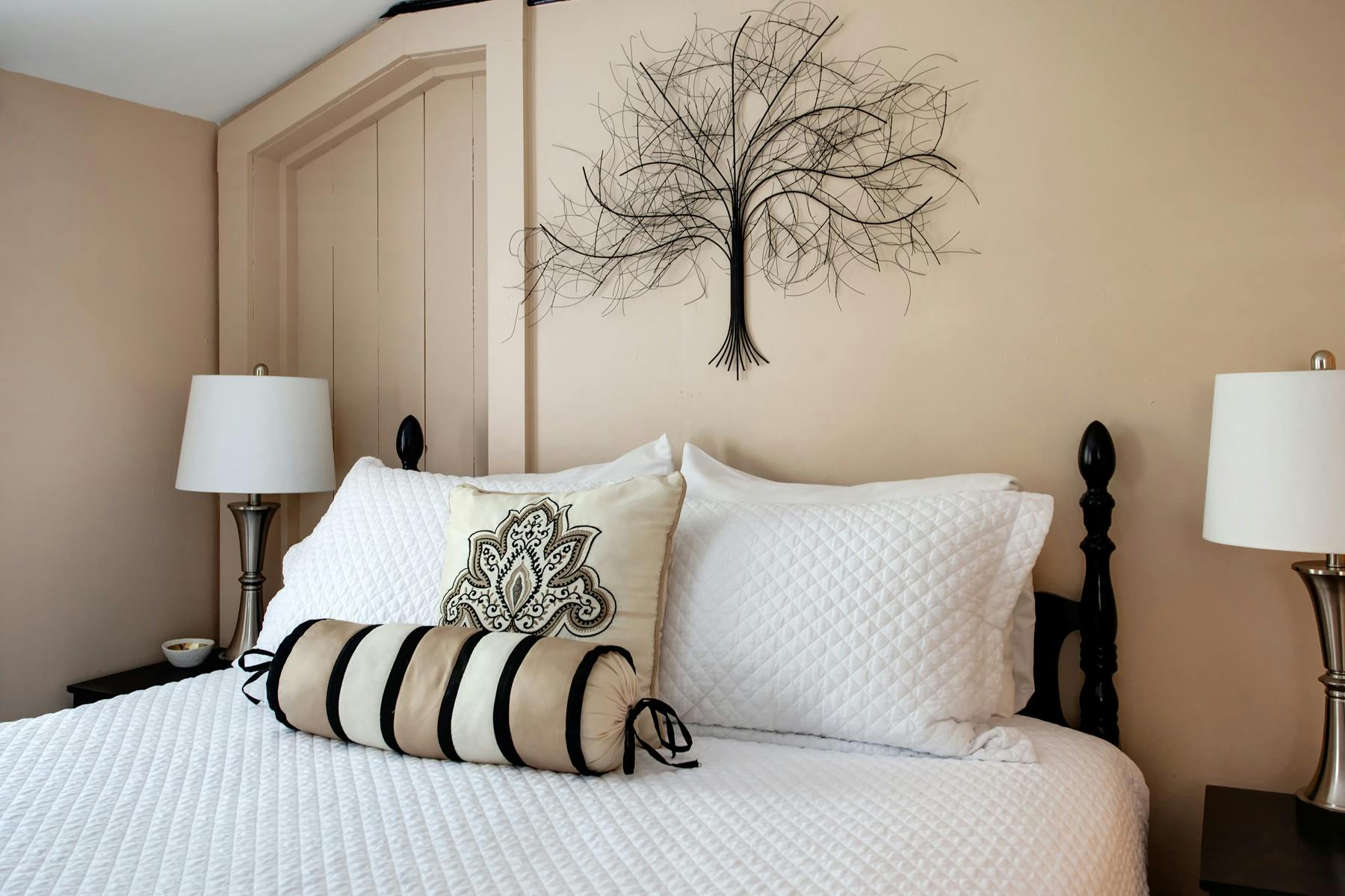 Close up of headboard of bed with decorative throw pillows, nightstands with matching lamps, and decorative tree on wall above