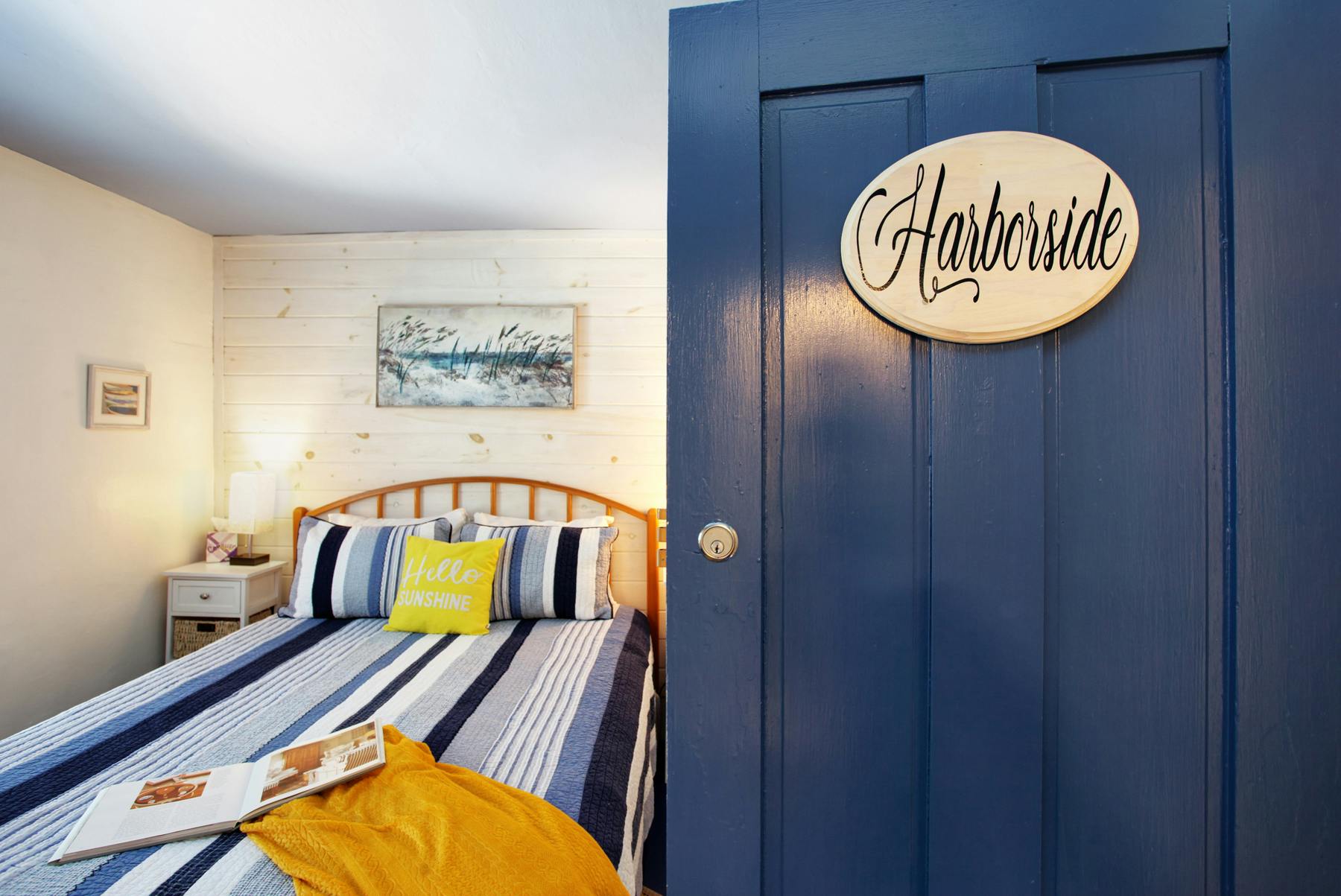 View of bedroom from doorway, bed with blue and white striped bedding with cheerful yellow accents, blue door with sign reading "Harborside"