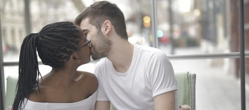 A young man and woman kissing while sitting in front of a window looking out to a blurred cityscape