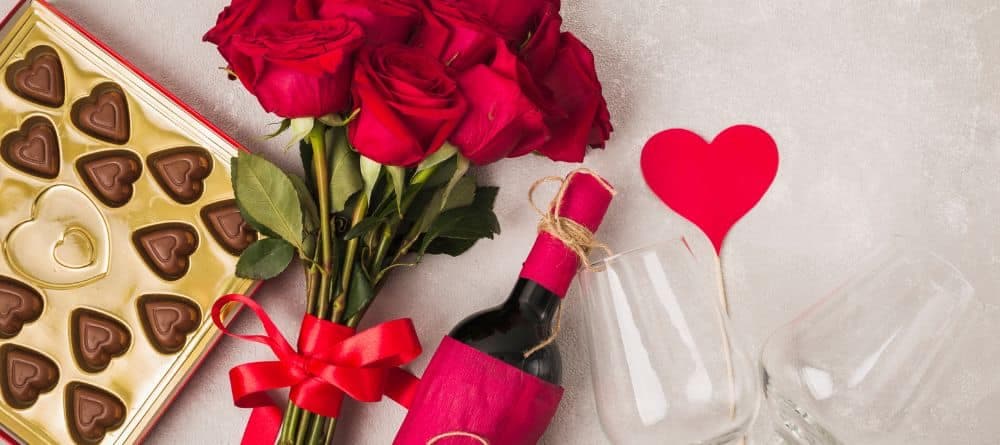 A box of heart-shaped chocolates, a bouquet of red roses, bottle of wine and two wine glasses.