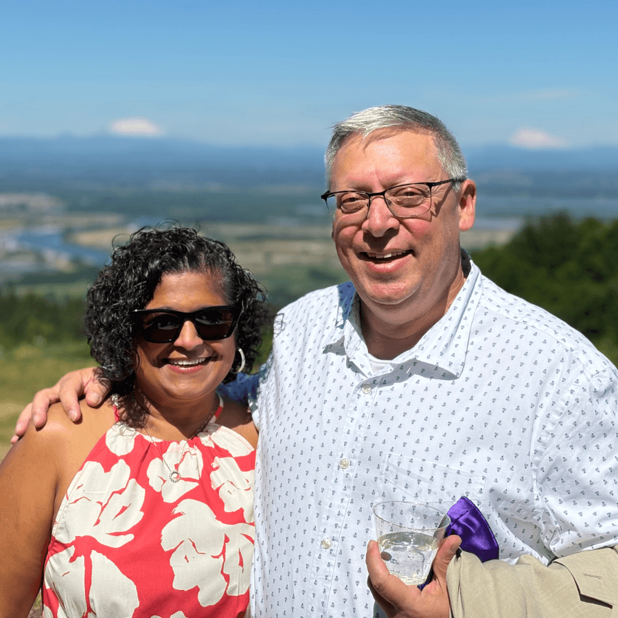 A smiling couple poses together outdoors with a scenic view in the background.